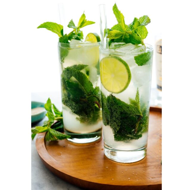 Two tall mojitos with fresh mint, lime wheels and ice on a wooden board