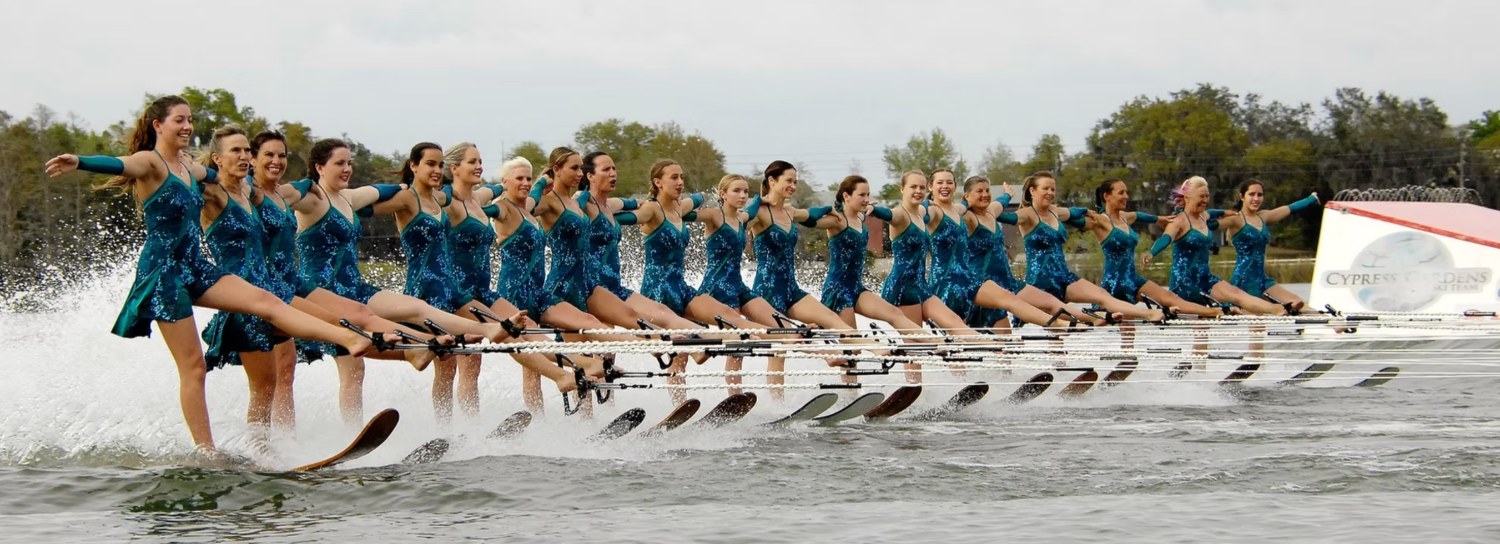 Long line of Cypress Gardens skiers in teal skimming in precision across the water