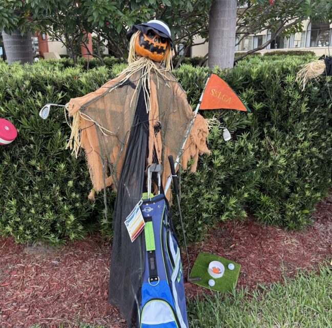 Golf-themed scarecrow built on a golf bag beside hedges at Sabal Springs