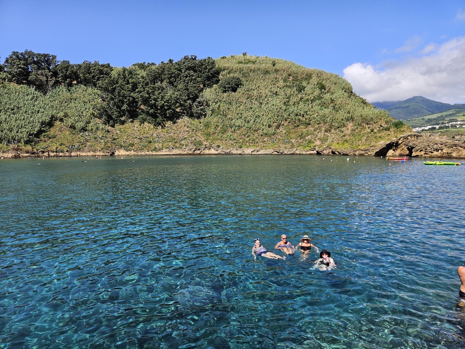 Swimmers in a clear turquoise volcanic pool below green cliffs.